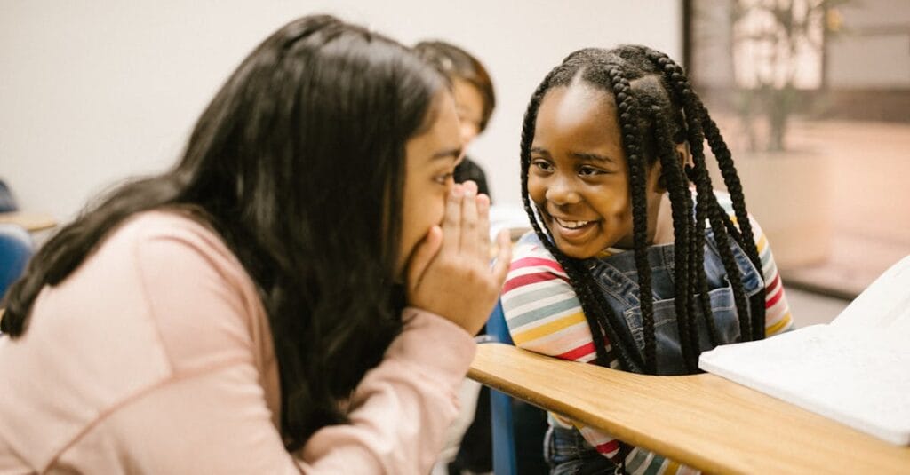 Two girls whisper and smile in a classroom, fostering friendship and joy.