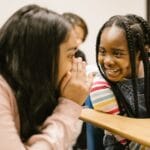 Two girls whisper and smile in a classroom, fostering friendship and joy.