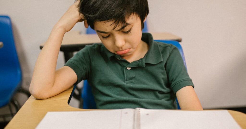 A young boy sits alone at a desk, looking sad and thoughtful in a classroom setting.