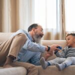A heartwarming moment between grandfather and grandson laughing and playing on a cozy sofa indoors.