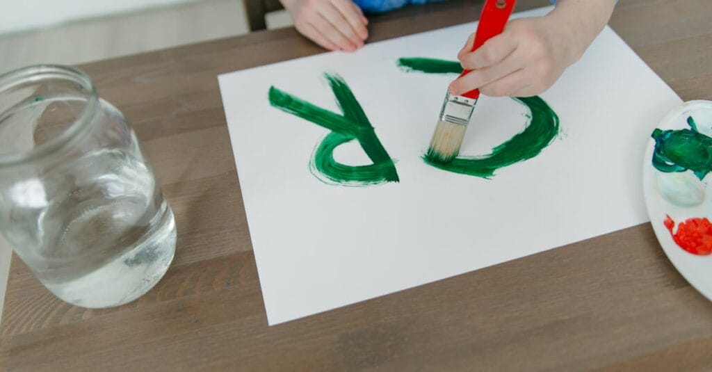 Child painting green letters on paper with a paintbrush. Creative indoor activity captured in detail.