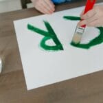 Child painting green letters on paper with a paintbrush. Creative indoor activity captured in detail.