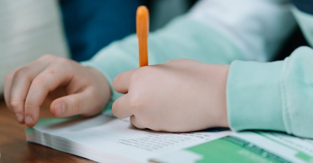 Close-up of a child's hands writing with an orange pen on a book indoors.