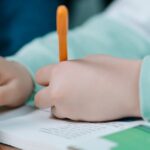 Close-up of a child's hands writing with an orange pen on a book indoors.