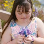 A young girl with Down syndrome enjoys nature, holding purple flowers by a wooden fence.