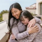 Mother and daughter with Down syndrome share a warm embrace outdoors on a sunny day.