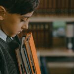 A young boy stands thoughtfully in a library, holding books. Education and solitude concept.