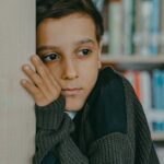 A contemplative child leans against a wall in a library, deep in thought.