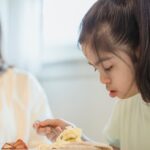 A young girl with Down syndrome enjoys breakfast with her mother in a cozy indoor setting.