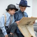 Two young boys in hats share a moment reading a newspaper, showcasing vintage style and curiosity.