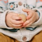 Close-up of a child's hands holding a wooden toy ball, wearing a cozy sweater indoors.