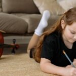 A child writing musical notes on sheet music while lying on a carpet indoors.