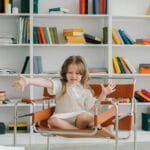 A young girl in a library sitting on a chair surrounded by colorful bookshelves.