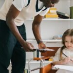 An adult male teacher assists a young girl in a classroom setting.