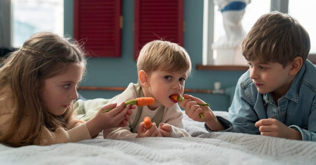 Kids lying on bed sharing fresh carrots in a cozy indoor setting.