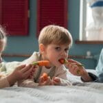Kids lying on bed sharing fresh carrots in a cozy indoor setting.