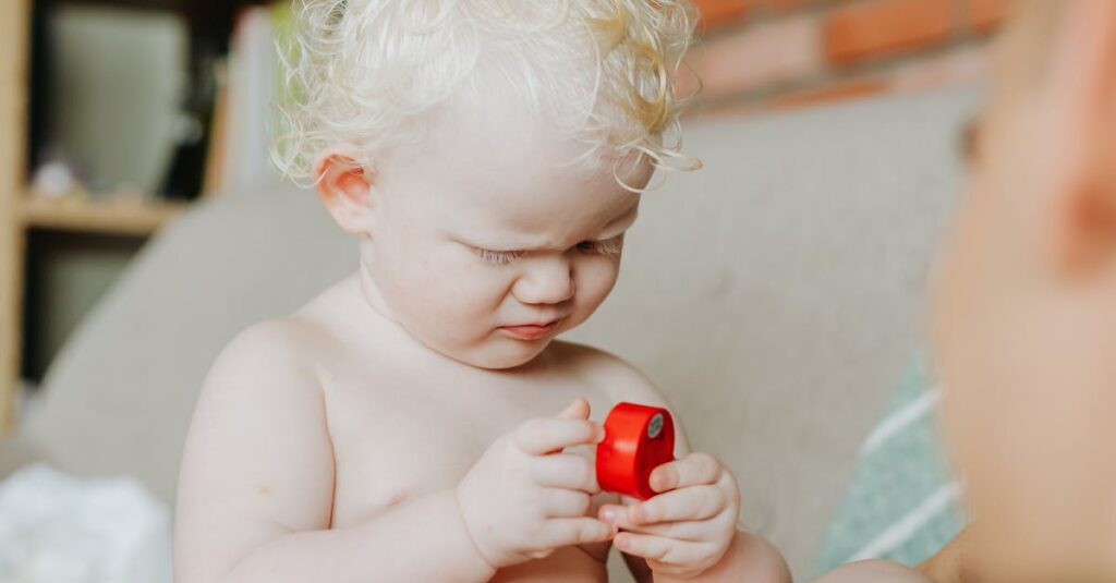 Cute baby with curly white hair plays with a red toy indoors, showcasing innocence and joy.