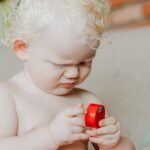 Cute baby with curly white hair plays with a red toy indoors, showcasing innocence and joy.