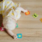 Cute baby in a polka dot dress crawls on wooden floor with colorful puzzle pieces.