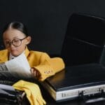 A child in eyeglasses and a yellow suit reads a newspaper at a desk, embodying a young journalist.