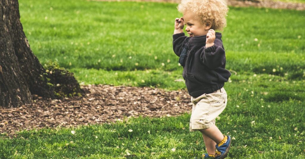 A happy toddler with curly hair joyfully runs on grass in a sunny park.