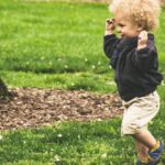 A happy toddler with curly hair joyfully runs on grass in a sunny park.