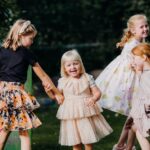 Four joyful children play in a garden, wearing colorful dresses and enjoying a sunny day.