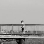 A young boy in a cap runs across a seaside bridge in a black and white setting.