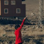 A child plays near a traditional house in İncesu, Türkiye, on a dry grassy field.