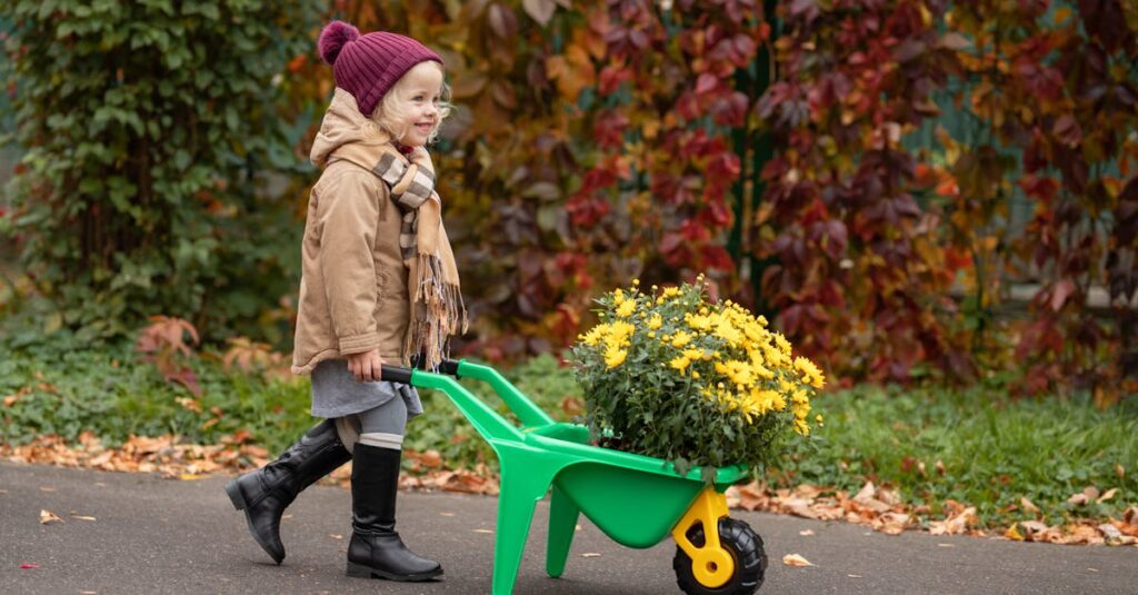 A young girl in autumn attire pushing a wheelbarrow filled with yellow flowers outdoors.