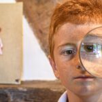 Redheaded boy holding a magnifying glass, amplifying one eye, indoors in Pont-l'Évêque, France.