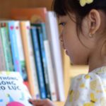 A young girl in a floral dress reading a book in front of a bookshelf.