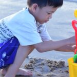 Young boy enjoys building sandcastles with colorful toys by the ocean shore on a sunny day.