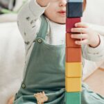 A cute child stacks colorful wooden blocks while playing indoors, surrounded by toys.