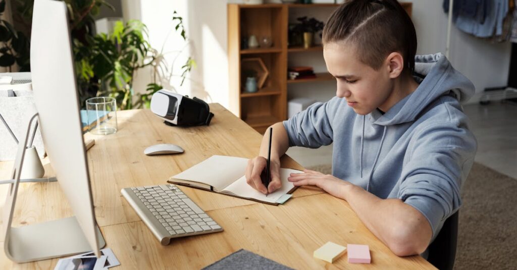 A teenage boy focuses on his studies at home, writing in a notebook during an online class.