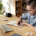 A teenage boy focuses on his studies at home, writing in a notebook during an online class.
