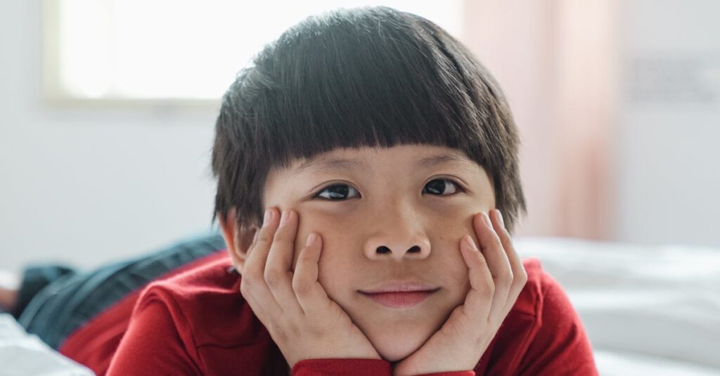 A happy child wearing a red shirt relaxes on a bed in a sunny, cozy bedroom, enjoying a peaceful morning.