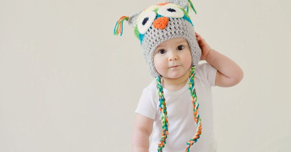 A cute baby wearing an owl hat standing in a basket, smiling and playful.