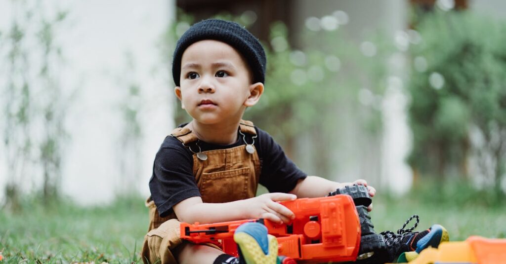 Cute boy in overalls playing with colorful toy trucks on the grass in a garden setting.