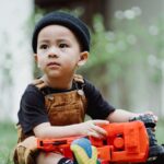 Cute boy in overalls playing with colorful toy trucks on the grass in a garden setting.