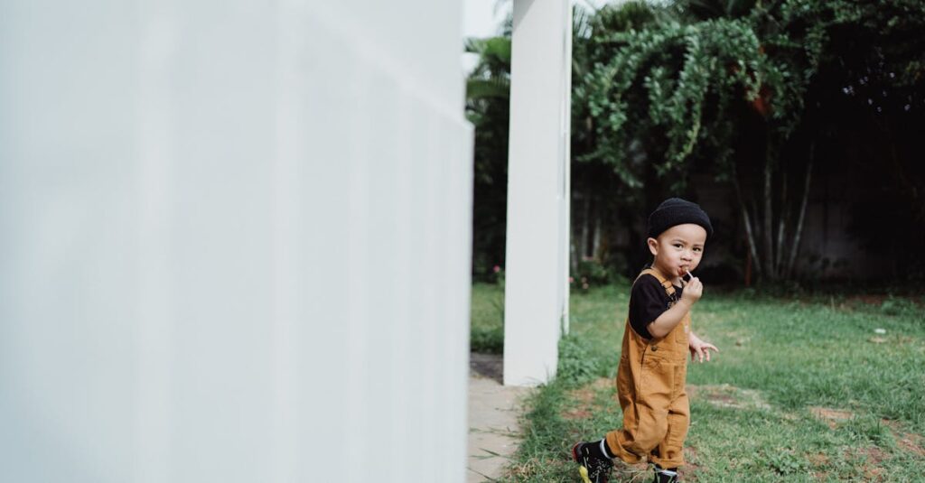 A young child in overalls enjoying a lollipop outdoors near a house.