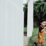 A young child in overalls enjoying a lollipop outdoors near a house.