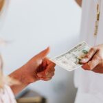 A woman in white hands a dollar bill to a child indoors, symbolizing financial exchange.