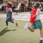 Two children enjoying playtime with capes on an outdoor playground, running on a sunny day.