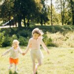 Two young girls holding hands and running on a grassy field in a sunny park setting.