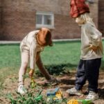 Unrecognizable little kids in casual outfits and brown hats playing with toys on backyard beside big tree