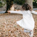 A young girl joyfully spins through fallen autumn leaves in a park, capturing the essence of carefree childhood.