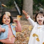 Two cheerful girls toss fall leaves joyfully in the park, capturing autumn fun.