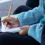 A young girl writing with a pencil while sitting outdoors in casual attire.