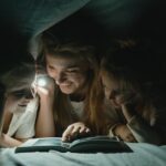 A mother reads a bedtime story to her children under the blanket with a flashlight, creating a warm, bonding moment.
